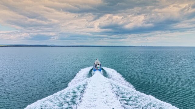 A boat moves through the water, creating waves behind it. The sky is mostly cloudy, and the horizon shows land far away. The scene captures a moment on the water.