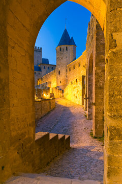Illuminated medieval city walls and towers frame a cobblestone street through a stone archway at dusk, offering a travel and tourism destination concept with copy space, Carcassonne, France.