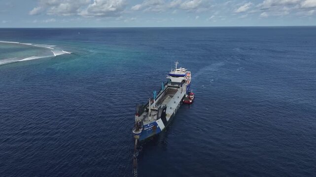 Aerial view of a large dredger ship navigating the deep blue waters near a coral reef under a cloudy sky in Gulhi, Kaafu Atoll, Maldives.