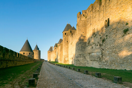 Historic medieval fortress walls and towers stand along a cobblestone path with green grass under a clear blue sky, offering a travel destination and copy space in Carcassonne, France.