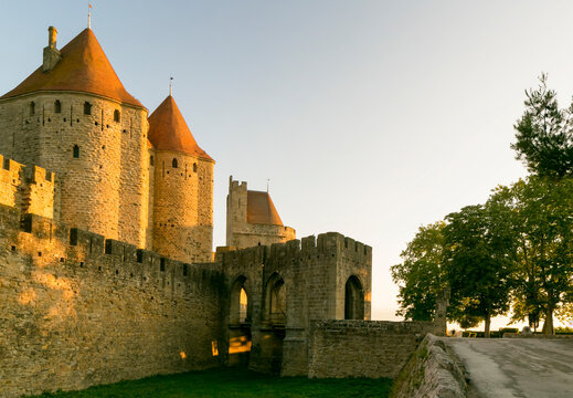 Carcassonne medieval fortress walls and towers glow under golden hour light against a clear sky, with green grass, a historic tourism destination in Carcassonne, Languedoc-Roussillon, France.