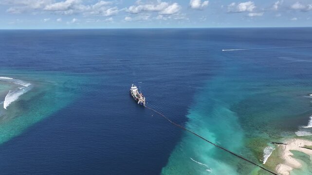 Aerial view of an industrial dredging ship with a long pipeline near a turquoise coral reef and deep blue ocean waters in Gulhi, Kaafu Atoll, Maldives.