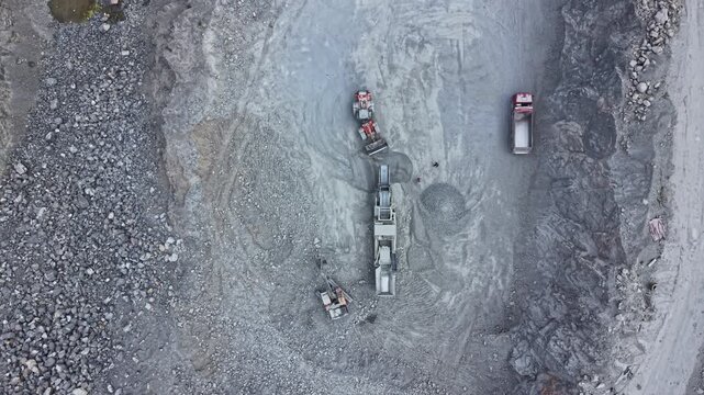Heavy machinery is working in a gravel pit to move rocks and materials. Trucks are lined up to transport the materials away from the site. This activity happens during daylight hours.