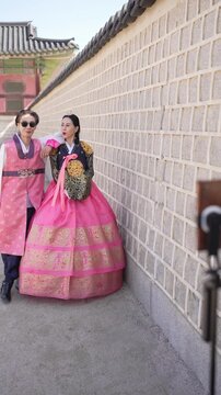 Two women in vibrant traditional Korean garments stand beside a stone wall. One wears a striking pink hanbok with gold patterns. The other dons a similar outfit, slightly darker in tone
