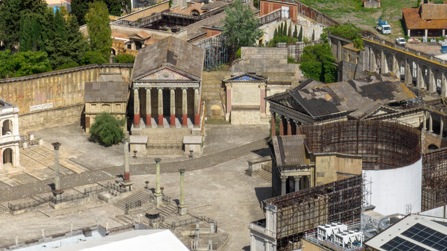 Aerial closeup of a detailed Roman film set in a studio backlot, featuring ancient temples, columns, and classical architecture amidst modern production equipment and infrastructure. View from above.