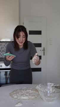Vertical screen woman checks artificial intelligence recipe on smartphone while failed dough sticks to her hands turning home cooking into stressful chaotic disaster