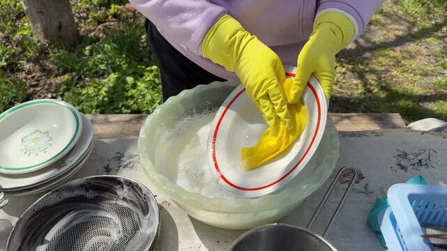 Both hands washing dishes outdoors A woman in yellow gloves washes plate with a yellow rag and a winterizing agent.