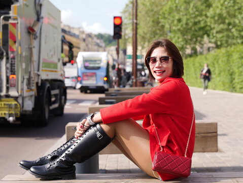 A woman wearing a red coat and tall black lace-up boots sits on a wooden bench as a city bus passes in the background