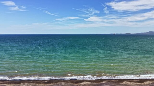 Waves flow gently onto a sandy beach where the ocean meets the land. The sky is blue with some clouds present. The location shows a distant shoreline and clear waters.