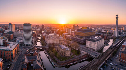 Obraz premium Panoramic view of a city skyline at sunset with the sun setting behind buildings and casting a warm glow.