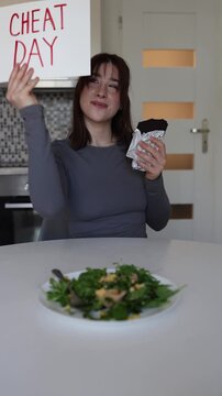 Vertical screen cheerful woman declares cheat day and chooses chocolate over healthy salad enjoying freedom from strict dieting and weight loss routine at home