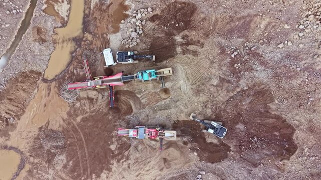 Heavy machinery works in a gravel pit. Trucks and machines transport material. The setting is near a stream where workers monitor the operation. The sun is shining in the afternoon.