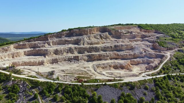 A large quarry with multiple layers of exposed rock. Dirt paths run through the area. The sky is clear and blue, indicating daytime with bright sunlight.