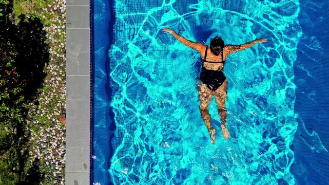 Aerial view of a woman in a black swimsuit swimming in a bright blue pool with sunlit ripples and caustic patterns Kuala Lumpur, Federal Territory of, Malaysia.