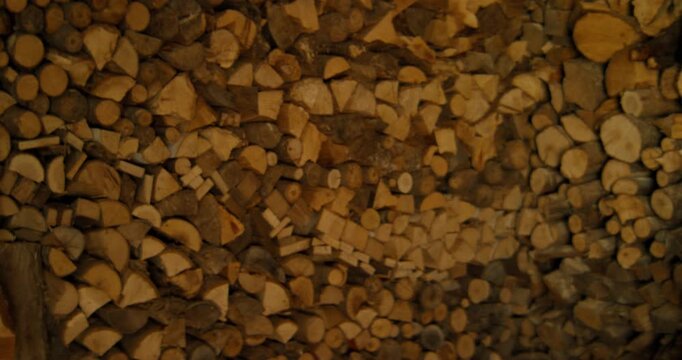 The camera tilts upward from a large pile of split firewood logs to reveal a weathered straw hat hanging from a wooden roof beam. Warm, natural light emphasizes the textures of the wood and straw.