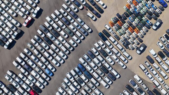 Aerial drone shot showing countless brand new cars neatly parked in rows, ready for shipment or sale at a large industrial distribution center or factory car park