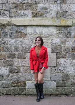 A woman in a red jacket and tall black lace-up boots stands smiling against a rustic stone wall