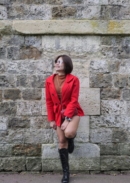 A woman wearing a red coat and sunglasses leans against a stone wall and looks upward