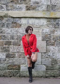 A woman wearing a red coat and sunglasses leans against a stone wall while laughing and looking to the side