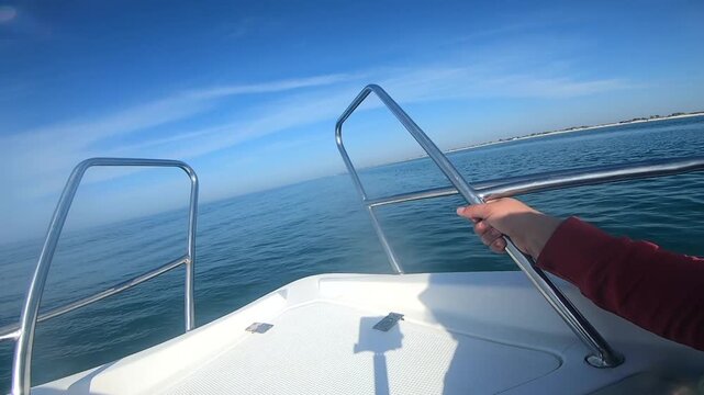 Woman holding onto metal handrails of motorboat sailing on sea on sunny summer day. Motorboat swimming fast, jumping on sea surface of water. View of side of boat. Leisure relax travel transportation