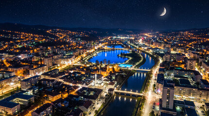 Obraz premium Panoramic aerial view of a city at night with illuminated buildings, winding river, and crescent moon in the starry sky.