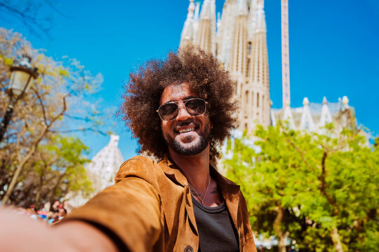 Smiling man with afro hairstyle enjoying a barcelona vacation, making a selfie during city sightseeing in front of sagrada familia.