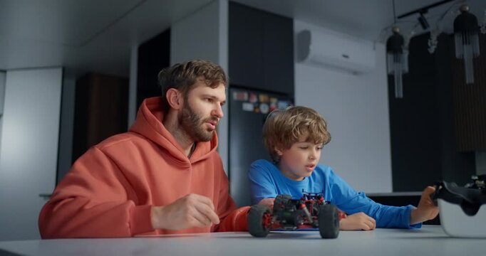 Father and young son assembling a remote control toy car together at a kitchen table, sharing a focused hands-on learning moment at home