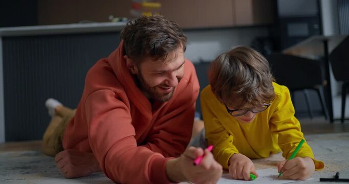 Father and young son lying on living room floor drawing together with colorful markers, sharing a warm creative family moment at home