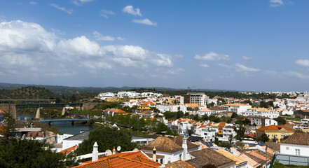 Fototapeta premium Tavira shows white houses and narrow streets from above, highlighting the tow's layout, greenery, and surrounding hills on a clear day in Algarve, Portugal