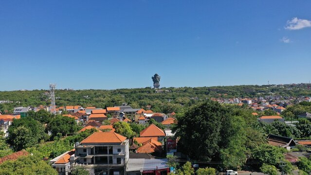 Panoramic aerial view of Jimbaran, Bali, Indonesia featuring tropical residential rooftops, lush green landscape, and the iconic Garuda Wisnu Kencana (GWK) statue under a clear blue sky. Beautiful urb