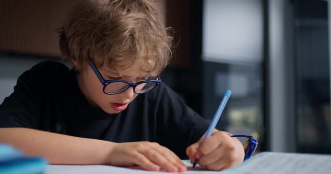 Closeup Of Boy Practicing Handwriting Skills. Young Boy With Blue Glasses Concentrates On Pen And Paper