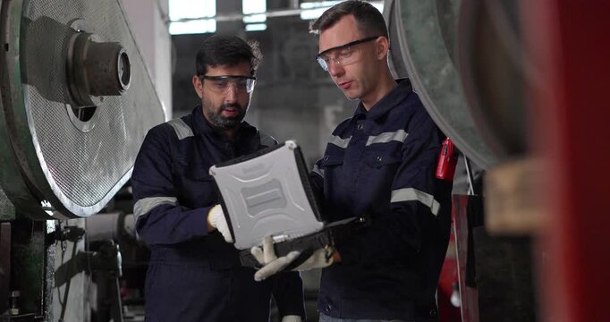 Two factory technicians review machine data on a laptop beside heavy industrial equipment in a traditional metal workshop, combining manual expertise, inspection, and modern diagnostics.
