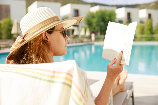 Woman reading book by poolside relaxing during summer vacation