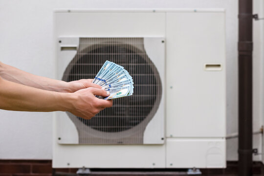 Heat Pump for Home Heating. Hand with Money Dollars on Heat Pump Background. Heat Pump Energy Savings for Efficient Home Heating System.