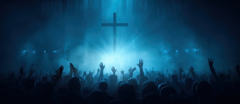 Brightly illuminated christian cross centered over crowd with raised hands during worship under blue light
