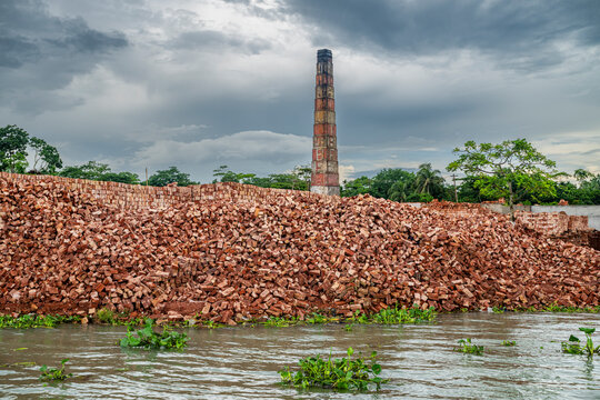 Brick factory operates on waterchannels in Barishal, Bangladesh under a cloudy sky with a large stack and piles of bricks