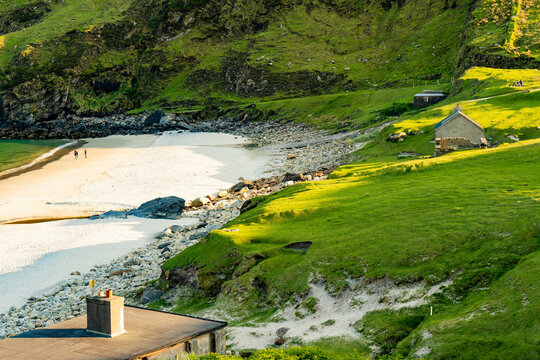 Keem Beach on Achill Island, County Mayo, Ireland, with calm blue water and a winding stream across the sand. This image shows the shoreline from land in warm light.