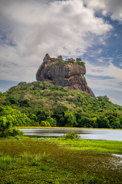 Rock fortress Sigiriya rises above water and lush greenery in central Sri Lanka at midday with a cloudy sky