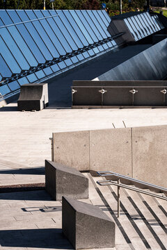 concrete plaza opens beneath steel canopy where stairs, ramp, railings, sunlight, shadows and urban architecture guide a civic exterior