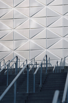 white wall rises beyond stairs as railings lead through geometric pattern shadows toward an urban entrance shaped by clean architecture