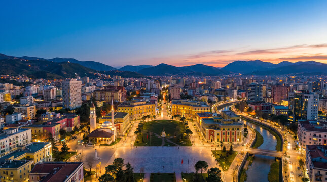 Aerial view of Skanderbeg Square in Tirana, Albania at dusk with city lights illuminating the buildings and surrounding mountains.