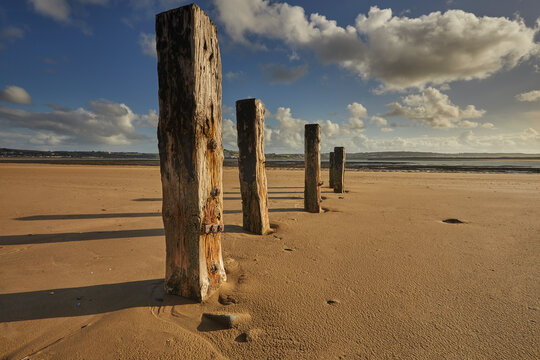 Old groynes stand on the beach at Crow Point in Devon under a clear sky with scattered clouds