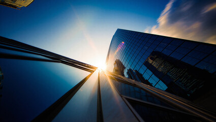 Low angle view of glass skyscraper reflecting bright sun flare against blue sky with high rise © Sadiya Digital
