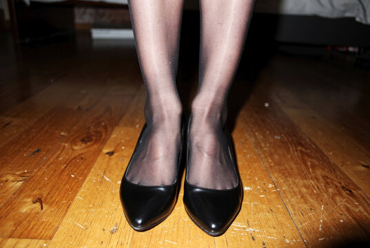 Elegant female legs in sheer black nylon stockings and classic black pointed-toe leather pumps standing on a polished wooden floor