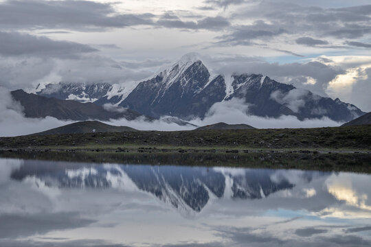 Snow-covered peak in Daxue Mountain Range reflects in Lenggacuo Lake at evening under soft light and clouds