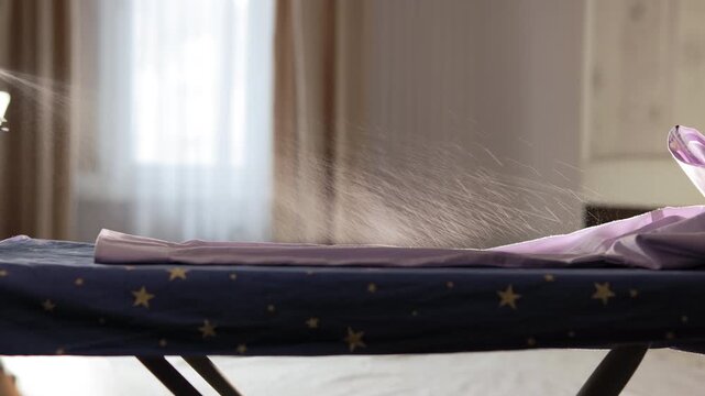 In living room, a man hand with iron sprays water onto a shirt lying on board