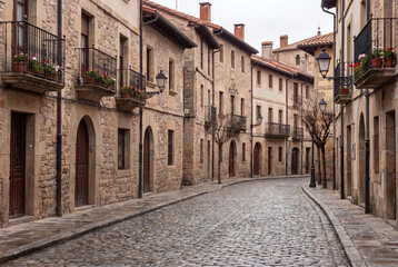 Obraz premium Charming empty cobblestone street winding through an old medieval European village featuring historic stone houses with balconies and street lamps.
