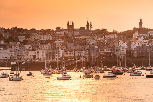 Sailboats are moored in St Peter Port Harbour during sunset with the skyline of Guernsey in the background