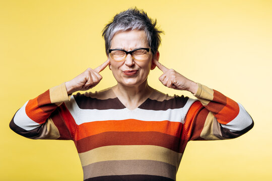 Mature woman covering her ears with fingers, expressing frustration with loud sound, standing on a yellow background
