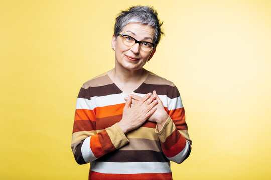 Grey-haired woman with short hair and glasses expressing sincere appreciation and empathy with hands placed over her chest on a yellow background